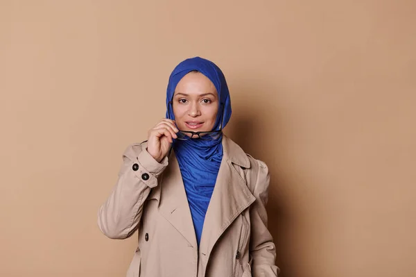 Attractive middle-aged Middle Eastern Muslim woman in blue hijab and casual beige coat, putting off her stylish spectacles, looking confidently at camera, isolated cream background. People Lifestyle