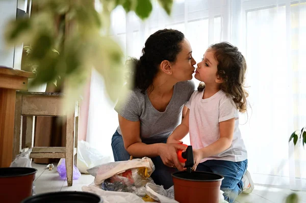 Loving mom kissing her daughter while taking care for houseplants. Little daughter learning gardening hobby in springtime, watering soil with plant seeds. Love and take care for planet from childhood