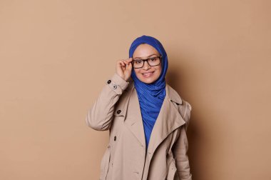 Çekici, şık, zarif, mavi tesettürlü, bej ceketli, şık gözlüklü, krem rengi arka planda izole bir şekilde kameraya gülümseyen Arap bir kadın. İnsanlar Moda