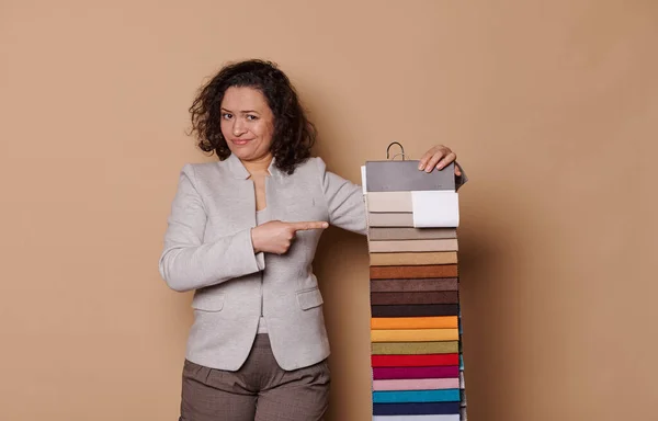 Hesitant Latin American curly haired woman in formal clothes, looking at camera, pointing at colorful fabric samples, having some difficulties on choosing sofa upholstery materials, beige background