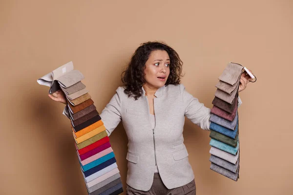 Hesitant Hispanic woman, interior designer, home decorator, or client in furniture store, holding fabric samples, having difficulties on choosing upholstery materials over isolated beige background.