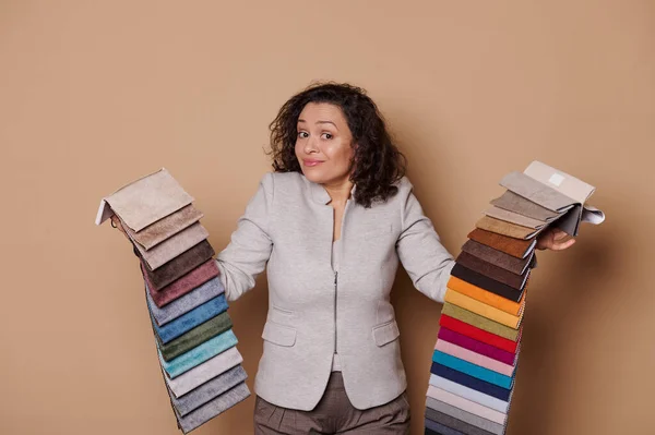 Charming multi-ethnic woman feels doubt while chooses upholstery textile, holds fabric samples on beige isolated background. Interior design. Home improvement, decoration, renovation
