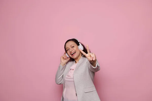 Cheerful woman in wireless headphones, showing peace sign at camera, enjoying new playlist, smiling and expressing happiness and positive emotions over isolated pink background. Copy advertising space