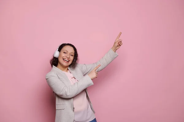 Lovely joyful positive ethnic woman pointing index fingers at copy ad space on pink background, enjoys new soundtrack and playlist, listens to music on wireless headphones, smiles looking at camera