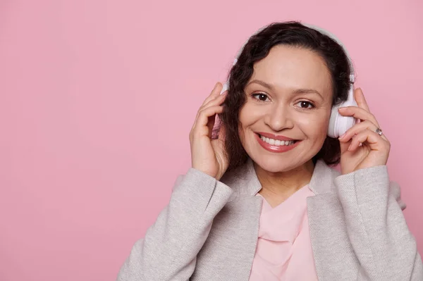 Headshot of a multi-ethnic middle-aged woman wearing wireless headphones, a music lover, enjoying new playlist and sound track, smiling with a beautiful toothy smile looking at camera, pink background