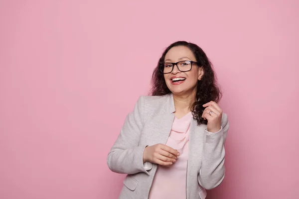 Happy multi-ethnic emotional middle-aged woman, wearing black trendy eyeglasses and elegant light gray suit, laughing looking at camera on isolated pink background. People, ophthalmology and fashion