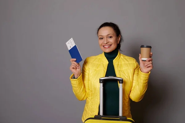 stock image Charming dark-haired multi-ethnic woman posing with cardboard cup of hot drink, suitcase and boarding pass, going for weekend getaway, cutely smiling looking at camera over gray isolated background