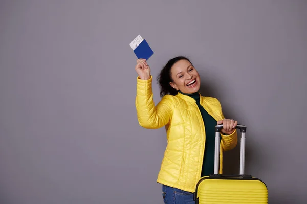 stock image Happy multi-ethnic woman showing her passport and air flight ticket, smiling looking at camera, expressing positive emotions standing near her yellow suitcase over isolated gray background. Copy space