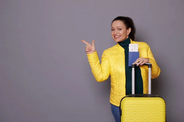 stock image Beautiful Latin American pregnant woman in yellow jacket, posing with boarding pass and color suitcase, smiling a toothy smile looking at camera, pointing at copy ad space on isolated gray background