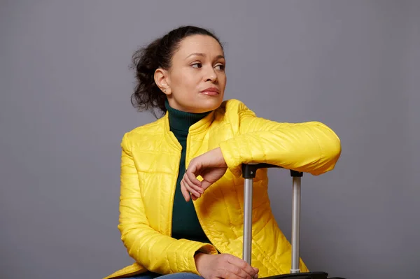 明るい黄色のジャケットで感情的な肖像多民族の動揺中年女性 離れて見て 悲しい感情を表現 広告のためのスペースと灰色の隔離された背景にスーツケースに座って — ストック写真