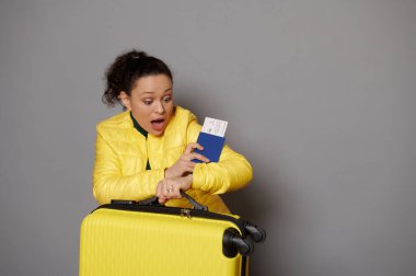 Sarı bavul ve uçak bileti taşıyan İspanyol genç bir turist. Kol saatine hayal kırıklığıyla bakıyor. Gri arka planda izole edilmiş uçağını kaçırdığı için şoka girdiğini söylüyor.