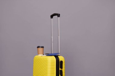 Tek kullanımlık eko kağıt bardak ve mavi pasaportla gri arka planda izole edilmiş parlak sarı bavul üzerinde izole edilmiş kahve ya da sıcak içeceğin reklam stüdyosu çekimi. Reklam alanını kopyala. Seyahat kavramı