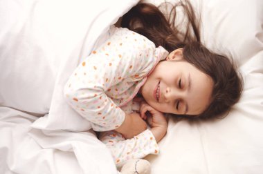 Beyaz pijama giymiş, beyaz pijama giymiş, açık renkli bir çocuk odasındaki rahat bir yatakta uyurken tatlı rüyalar gören beyaz, güzel bir kız çocuğunun manzarası.