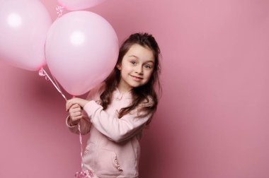 Beyaz, sevimli, küçük bir kız çocuğu şişme pembe pastel balonlardan seçiyor, kameraya dikkatlice bakıyor, pembe renkli arka planda izole edilmiş. Çocuklar işte. Mutlu bir çocukluk. Duygular. Kavramlar