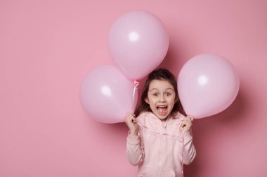Pembe kapüşonlu, elinde pembe pastel balonlar tutan, şaşırdığını ve şaşırdığını ifade eden küçük kız çocuğu, izole edilmiş pembe arka planda ağzıyla poz veriyor ve metin reklamları için reklam alanı yaratıyor.