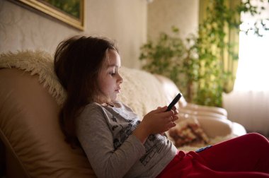 Sevimli küçük kız çevrimiçi film ya da video seyrediyor, evdeki kanepede otururken cep telefonuyla sanal oyun oynuyor. Çocukluktaki alet bağımlılığı sorunu. Çocuklar ve dijital teknolojiler