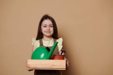 5 yaşında güzel bir kız çocuğu bahçe işleri hobisi yapıyor, kameraya gülümsüyor, bahçe aletleri ve sulama kutusuyla ahşap bir sandık taşıyor, ekime ve dikmeye hazır, izole edilmiş bej arka plan.