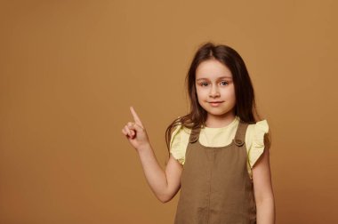 Birinci sınıf öğrencisi ya da birinci sınıf öğrencisi, şık haki elbiseli, kameraya bakıp gülümseyen ve izole edilmiş bej arka planda işaret parmağıyla işaret eden kız çocuğu. İlkokul öğrencisi. Çocuklar