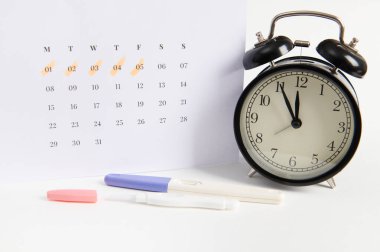 Pozitif mürekkep püskürtme hamilelik testi, siyah retro alarm saati ve son adet tarihlerinin yazılı olduğu beyaz takvim. Kadın sağlığı, yumurtlama, doğurganlık, hamilelik planlaması ve annelik. Reklam alanı