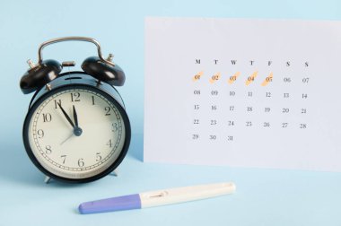 Pozitif mürekkep püskürtme hamilelik testi, siyah retro alarm saati ve mavi arka planda son adet tarihlerinin yazılı olduğu beyaz takvim. Kadın sağlığı, yumurtlama, doğurganlık, hamilelik planı