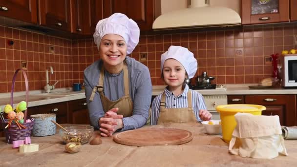 シェフの帽子とエプロンの陽気な母親と娘は カメラを見て 自宅のキッチンのインテリアに立って笑顔で イースターのパネトーネのためのペストリー生地を準備します キリスト教の伝統と祝日 — ストック動画