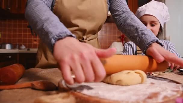 ベージュエプロンで主婦の女性のクローズアップ 圧延ピンで生地をロールアウト 彼女の娘は料理を勉強している ママと子供は一緒に家のキッチンでジンジャーブレッドクッキーを調理します — ストック動画