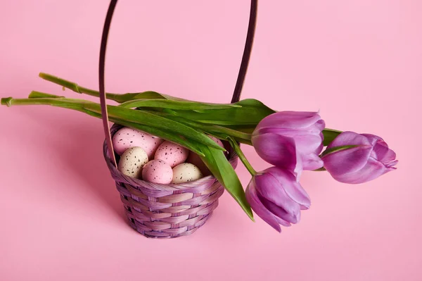 Purple basket filled with Easter eggs and bouquet of tulips on pink background. Copy ad space. Easter celebration concept, a religious holiday in christianity symbolizing Jesus Christ resurrection