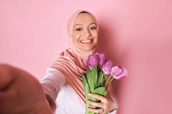 ピンクのヒジャーブで陽気な若い中東イスラム教徒の女性 美しいおもちゃの笑顔で笑顔ながら 彼女の外側の手でスマートフォンで自画自賛します ピンクの背景にチューリップの花束とポーズ — ストック写真