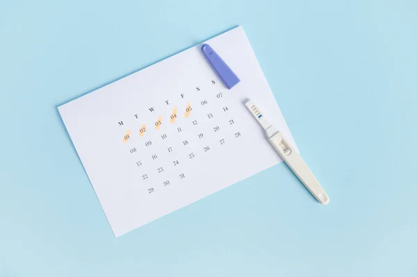 Top view of pregnancy test with two bars on white calendar with the menstruation last days marked, isolated on blue background. The concept of calculating ovulation, planning and diagnosis pregnancy