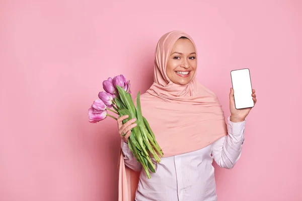 stock image Gorgeous Middle-Eastern Muslim woman in pink hijab, showing at camera a smartphone with white blank digital screen with ad space for mobile application, posing with tulips on isolated pink background