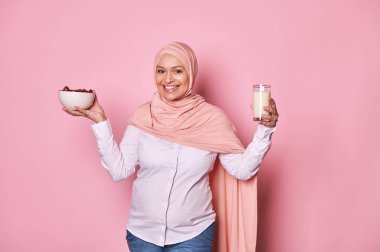 Pembe tesettürlü, güzel bir Orta Doğulu Müslüman kadın, Ramazan 'da sağlıklı bir kahvaltı hazırlıyor. Bir kase dolusu tatlı hurma ve sütlü, neşeli, dişlek bir gülümseme, pembe bir arka planda izole edilmiş.