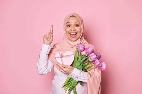 幸せな妊娠中の若いアラブイスラム教徒の女性でピンクヒジャーブ チューリップやギフトボックスの花束を保持します コピー広告スペースで指を指して 陽気に笑顔 驚きを表現します 孤立ピンクの背景 — ストック写真