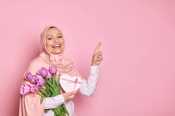 ピンクのヒジャーブに覆われた頭を持つ幸せなスタイリッシュなアラブのイスラム教徒の女性 母の日のための紫色のチューリップとギフトボックスの花束を保持 コピースペースで指をポイント カメラを見て笑顔 ピンクに隔離された — ストック写真