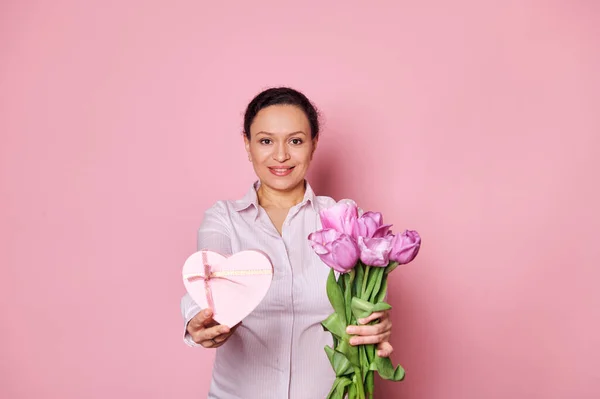 stock image Middle-aged ethnic pretty woman holding out at camera a beautiful bouquet of tulips and a heart shaped gift box, greeting with Mothers, International Womens or Saint Valentines Day, isolated on pink