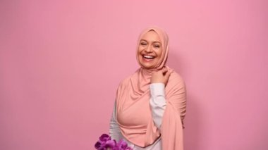 Pembe tesettürlü güzel Arap kadın, özel şenlik için mor laleler tutuyor, pembe arka planda izole edilmiş kameraya bakıp gülümsüyor. Uluslararası Kadınlar ve Anneler Günü