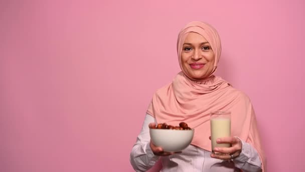用粉色头巾微笑的穆斯林女人 说拉马丹 卡里姆 同时给你一杯健康的牛奶和一碗甜枣子 在宗教禁食后享用完美的早餐 开斋节 伊斯兰文化 — 图库视频影像