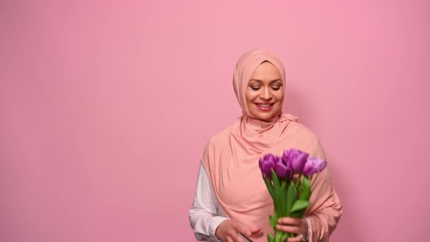 ピンクのヒジャーブの美しい中年アラブイスラム教徒の女性は 紫のチューリップの束を嗅ぎ 幸福と肯定的な感情を表現し 笑顔とカメラを見て 国際女性母の日 — ストック動画
