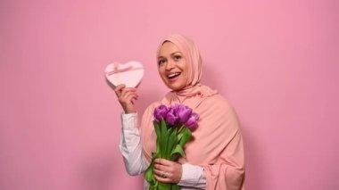 Pembe tesettürlü mutlu Ortadoğulu Müslüman kadın, pembe kalp şeklinde hediye kutusunu sallıyor, içinde ne olduğunu tahmin etmeye çalışıyor, mor lale buketi tutuyor, gülümsüyor, pembe arka plandaki kameraya bakıyor.