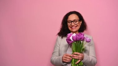 Orta yaşlı, çok ırklı, gözlüklü güzel bir kadın. Bir yandan mutlu olurken bir yandan da tebrik kartlarıyla bir buket lale alıyor. Sevimli bir şekilde gülümsüyor.