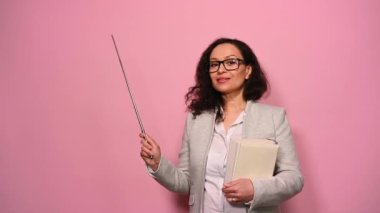 Elinde ders kitapları olan, gülümseyen, kameraya bakan ve izole edilmiş pembe arka plandaki fotokopi alanını işaret eden çok ırklı pozitif kadın öğretmen. Uluslararası Öğretmenler Günü.