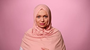 Kafası pembe tesettüre bulanmış, arka planda izole bir şekilde kameraya güvenle bakan orta yaşlı, güzel, Ortadoğulu Müslüman bir kadının yakın plan portresi. İslam Arap kadın modası