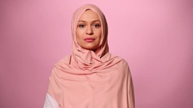 Kendine güvenen, orta yaşlı, pembe tesettürlü güzel bir Arap kadının yakından çekilmiş portresi. Cilveli bir şekilde gülümseyen, pembe arka planda izole edilmiş kameraya bakan. Orta Doğulu kadın modası. İslam kültürü