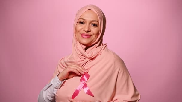 穿着粉色头巾的快乐的中年穆斯林妇女 双手环绕着缎带 对着镜头笑着 乳腺癌宣传运动 粉红月10月1日 孤立的色彩背景 — 图库视频影像