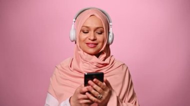 Çekici, mutlu, pembe tesettürlü Müslüman bir kadın, bir müzik aşığı, kablosuz kulaklıklarda yeni soundtrack dinleyen, akıllı telefondan sarkan çalma listesi, gülümseyen, dans eden, soyutlanmış pembe arka plan