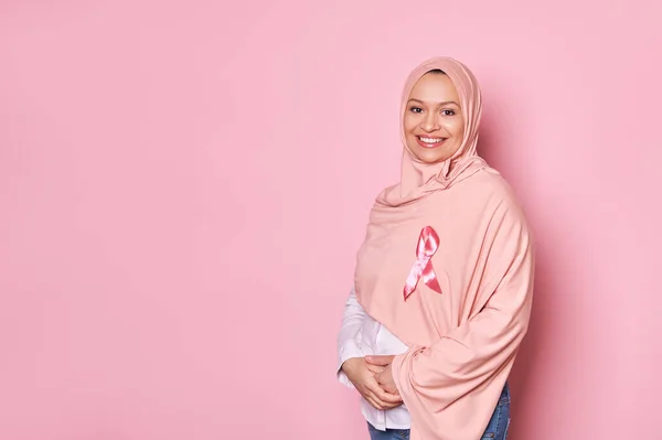 stock image Smiling Middle-Eastern Muslim pregnant woman in pink hijab, wearing satin ribbon, emblem of fighting breast cancer and showing support for cancer survivors and patients, isolated over pink background