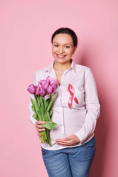 Smiling pleasant pregnant woman with pink satin ribbon, holding bouquet of purple tulips, supporting cancer patients, isolated pink background. October pink. Awareness campaign fighting breast cancer
