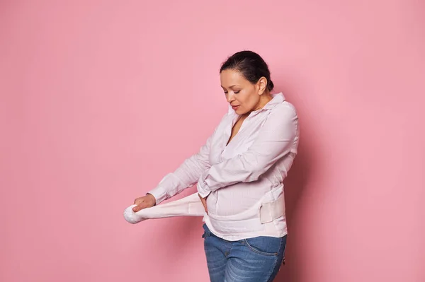 Pregnant woman putting on supporting bandage to reduce backache during pregnancy, isolated over pink background with copy advertising space. Orthopedic abdominal support belt concept. Pregnancy.