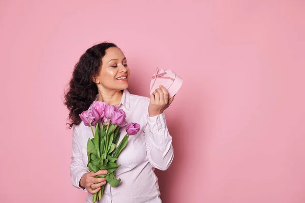 stock image Happy Latin American middle-aged woman smiles, rejoicing at cute present in a pink heart shaped gift box and a bouquet of purple tulips for Mothers or International Womens Day, isolated on pink