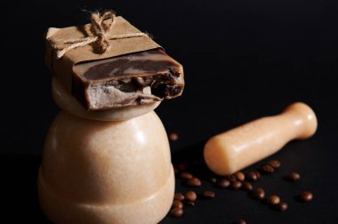 Soğuk baskılı ev yapımı organik sabun üzerine seçici bir odak noktası, bej mermer havan, siyah arka planda dağınık kahve çekirdekleri ve tokmağı ile izole edilmiş vücut fırçası. Spa, hijyen, vücut bakımı