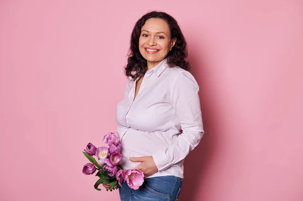 stock image Portrait of multi-ethnic positive pregnant woman, touching belly, smiling looking at camera, posing with bunch of tulips on isolated pink background. Maternity. Pregnancy. Happy Mothers Day concept